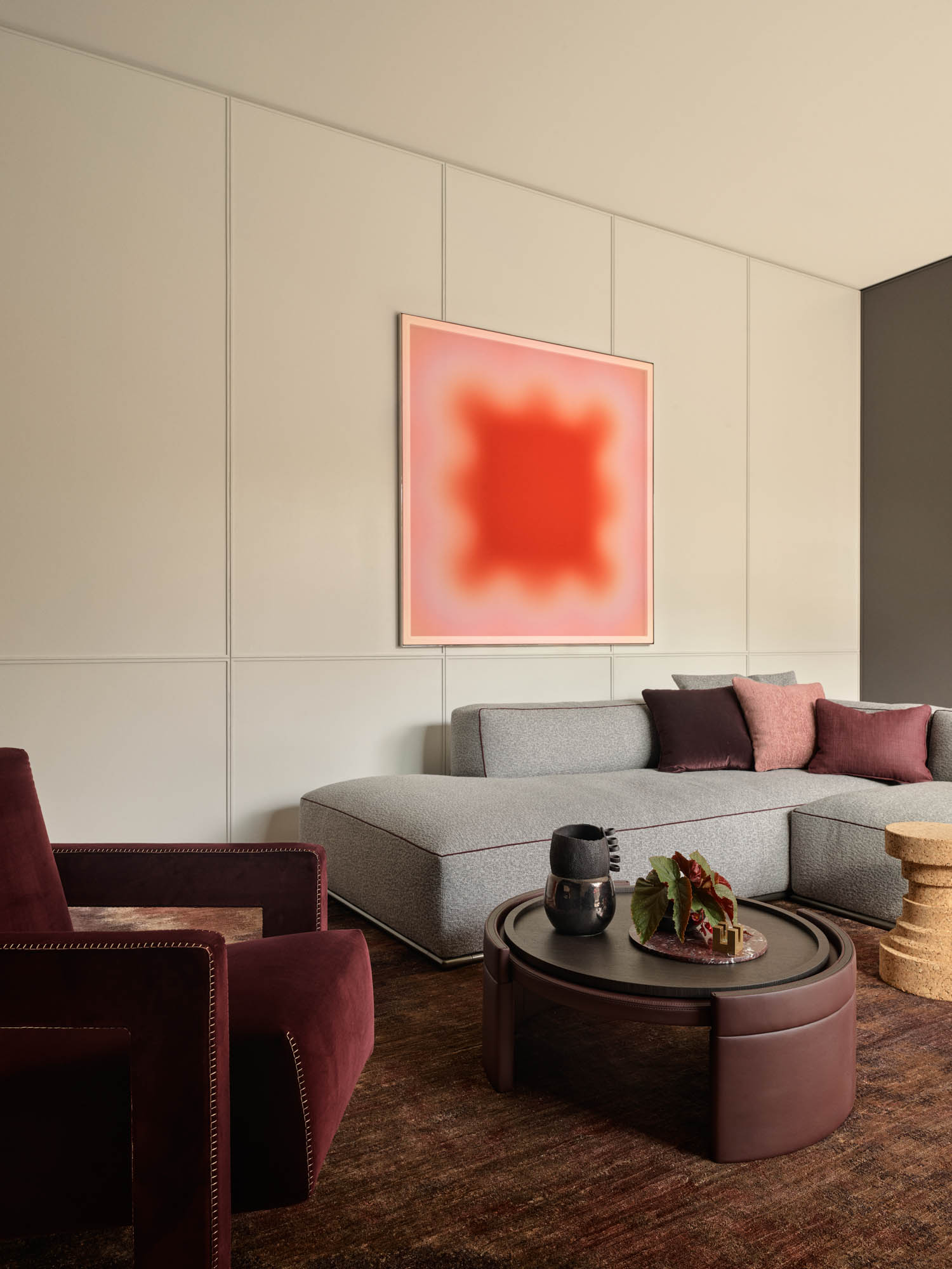 Modern living room with a gray sectional sofa, burgundy armchair, round coffee table, and abstract red artwork on the wall.