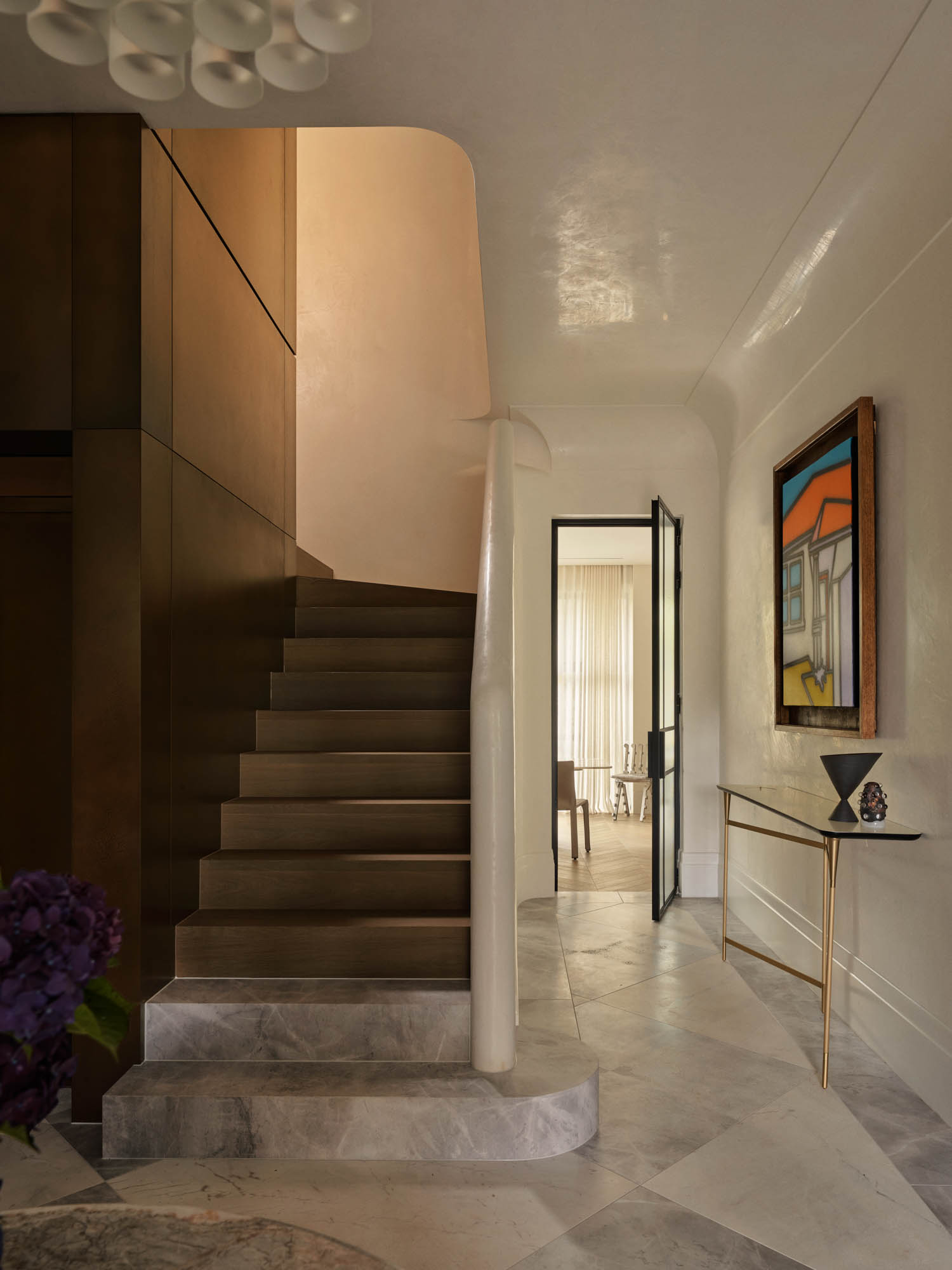 A wooden staircase with a curved white railing leads upstairs. A modern painting and slim console table are on the right wall. Marble floors and neutral walls complete the entryway.