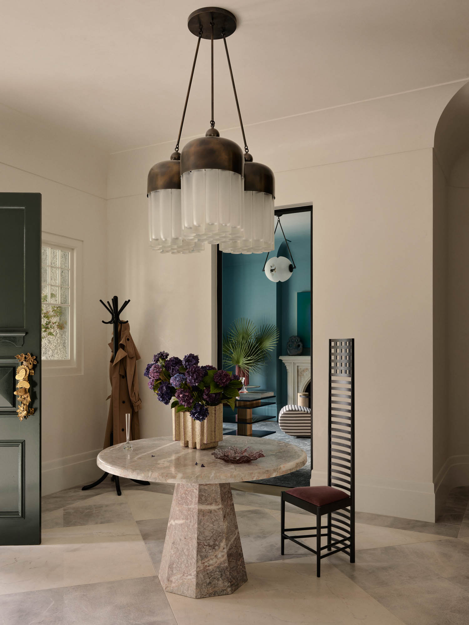 A round stone table with a floral arrangement and one high-backed chair stands under a modern chandelier in a neutral-colored entryway. A coat rack and open door are visible.