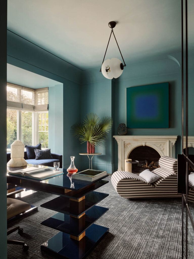 Modern living room with teal walls, striped chaise lounge, geometric black table, abstract art above a white fireplace, and a large palm leaf in a vase by the window.