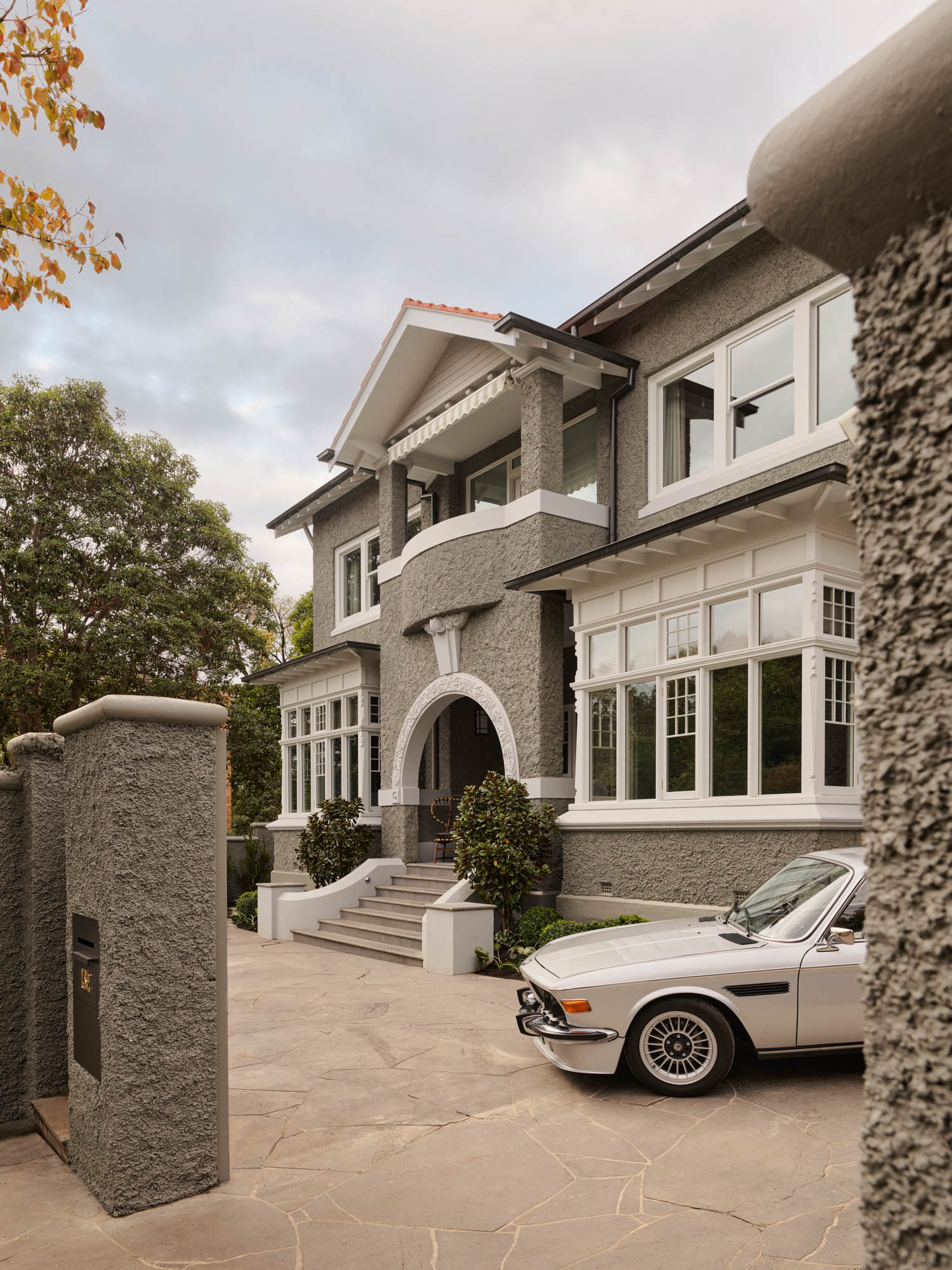 Gray stucco two-story house with large white-framed windows, arched entryway, and a vintage white car parked in the driveway.
