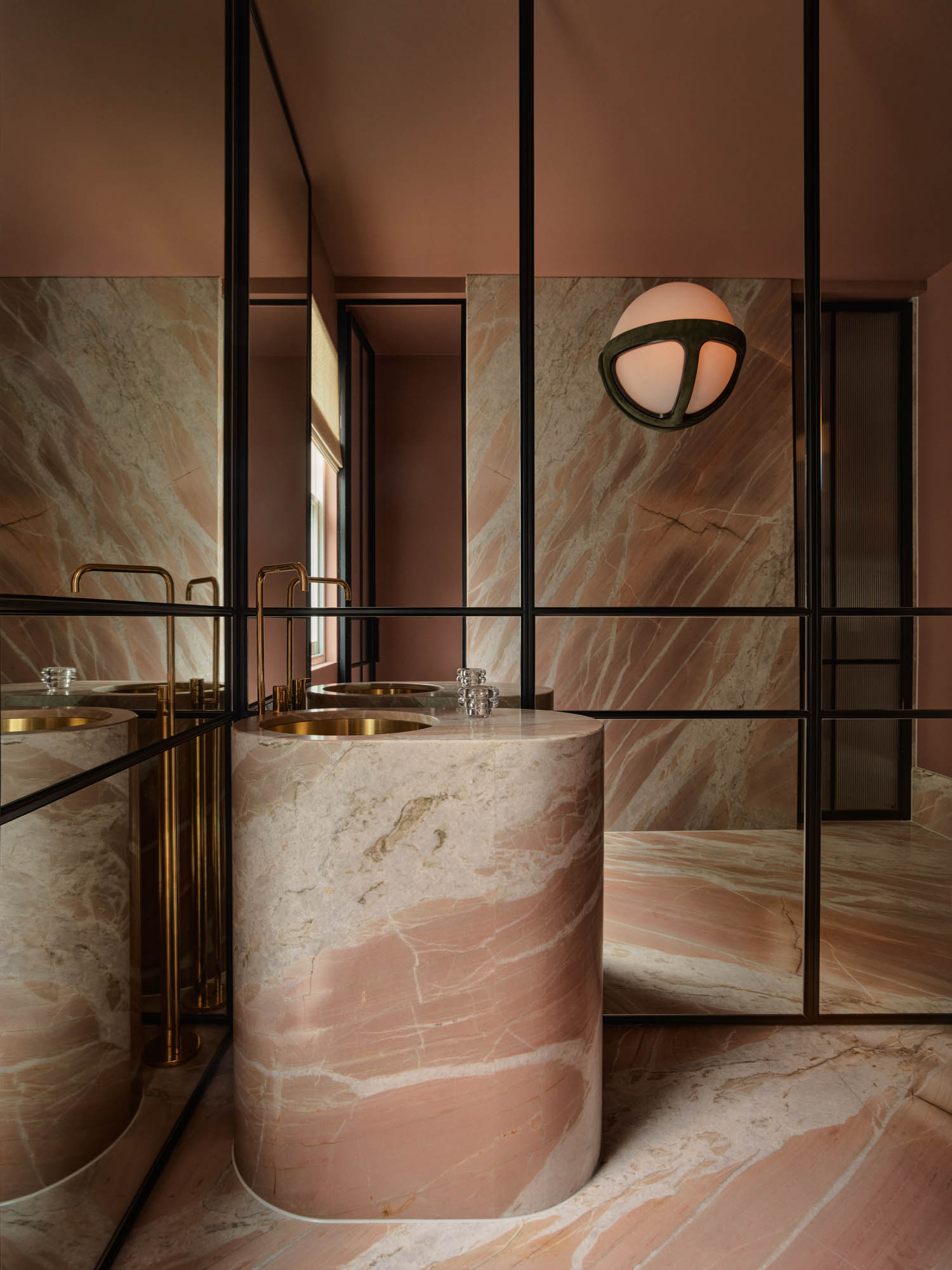 Modern bathroom with pink marble walls and cylindrical marble sink, gold faucet, glass tumbler, and round wall light fixture; mirrors reflect the space.