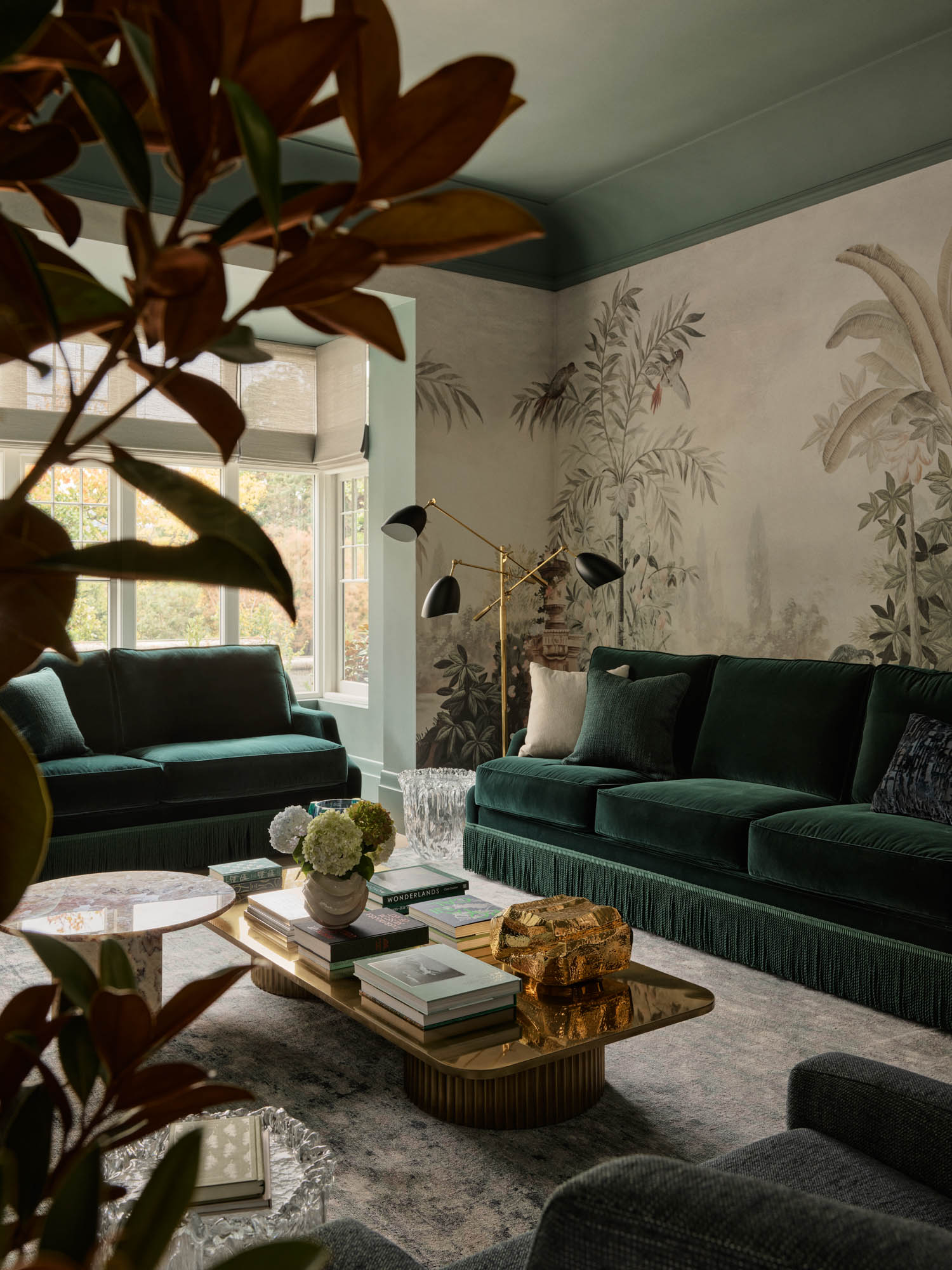 Elegant living room with green velvet sofas, gold coffee table, floral wallpaper, large window, and modern black floor lamp; leafy plant in the foreground.
