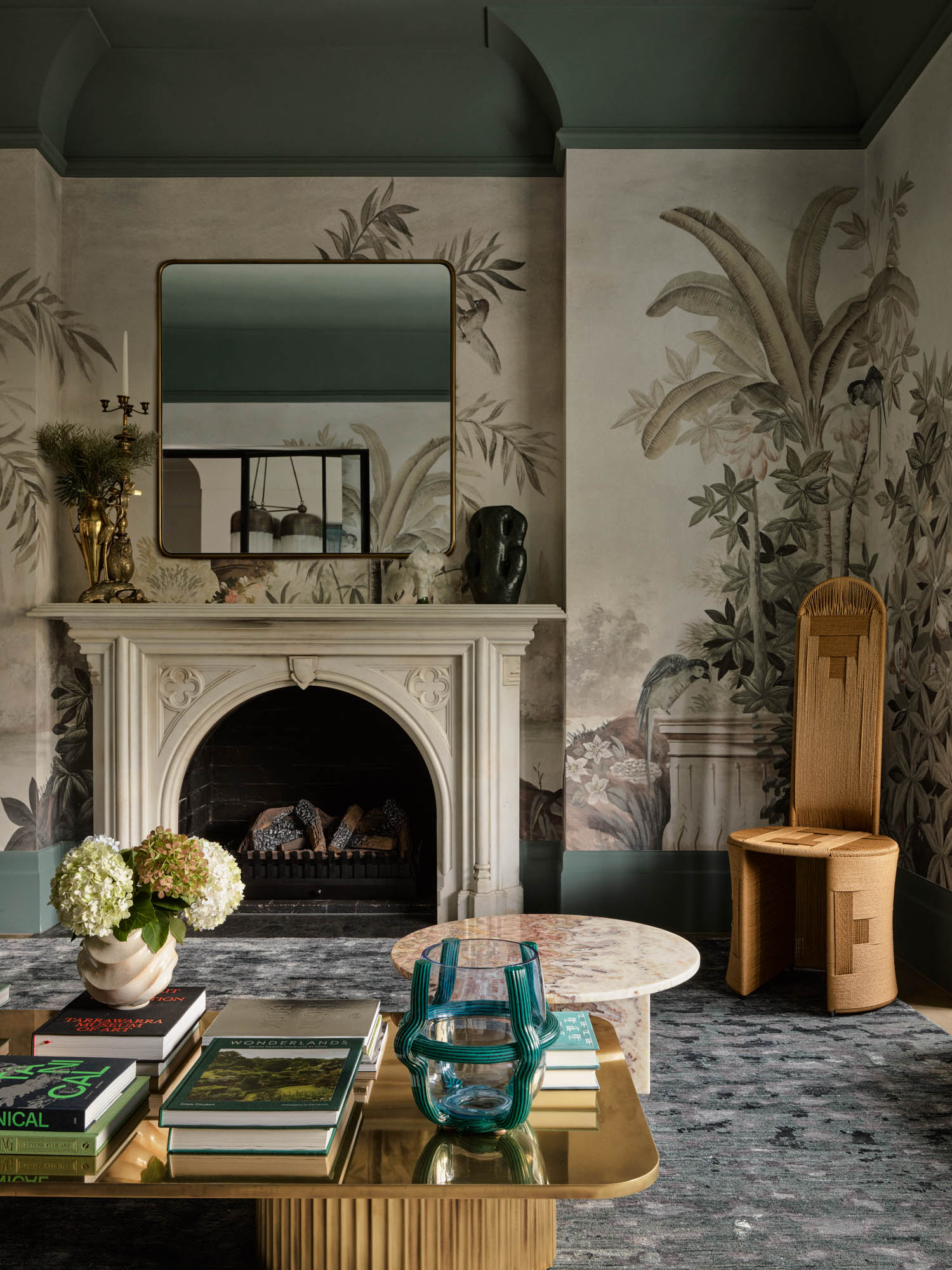 Elegant living room with ornate fireplace, botanical wallpaper, tall sculptural chair, coffee table with books and flowers, and a mirror above the mantel.