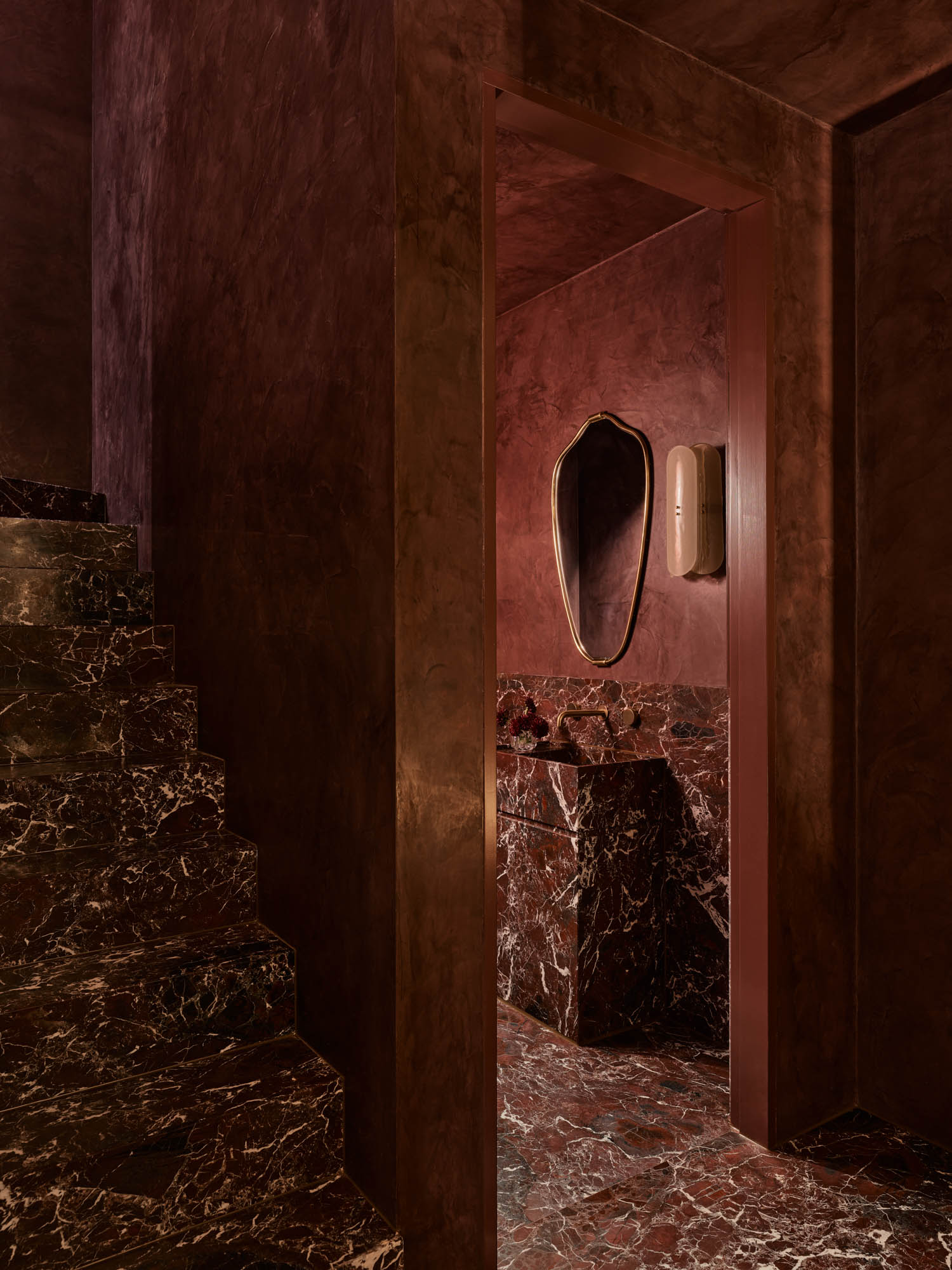 A marble staircase leads to a restroom with dark red marble walls, a geometric mirror, and a soap dispenser, all in warm ambient lighting.