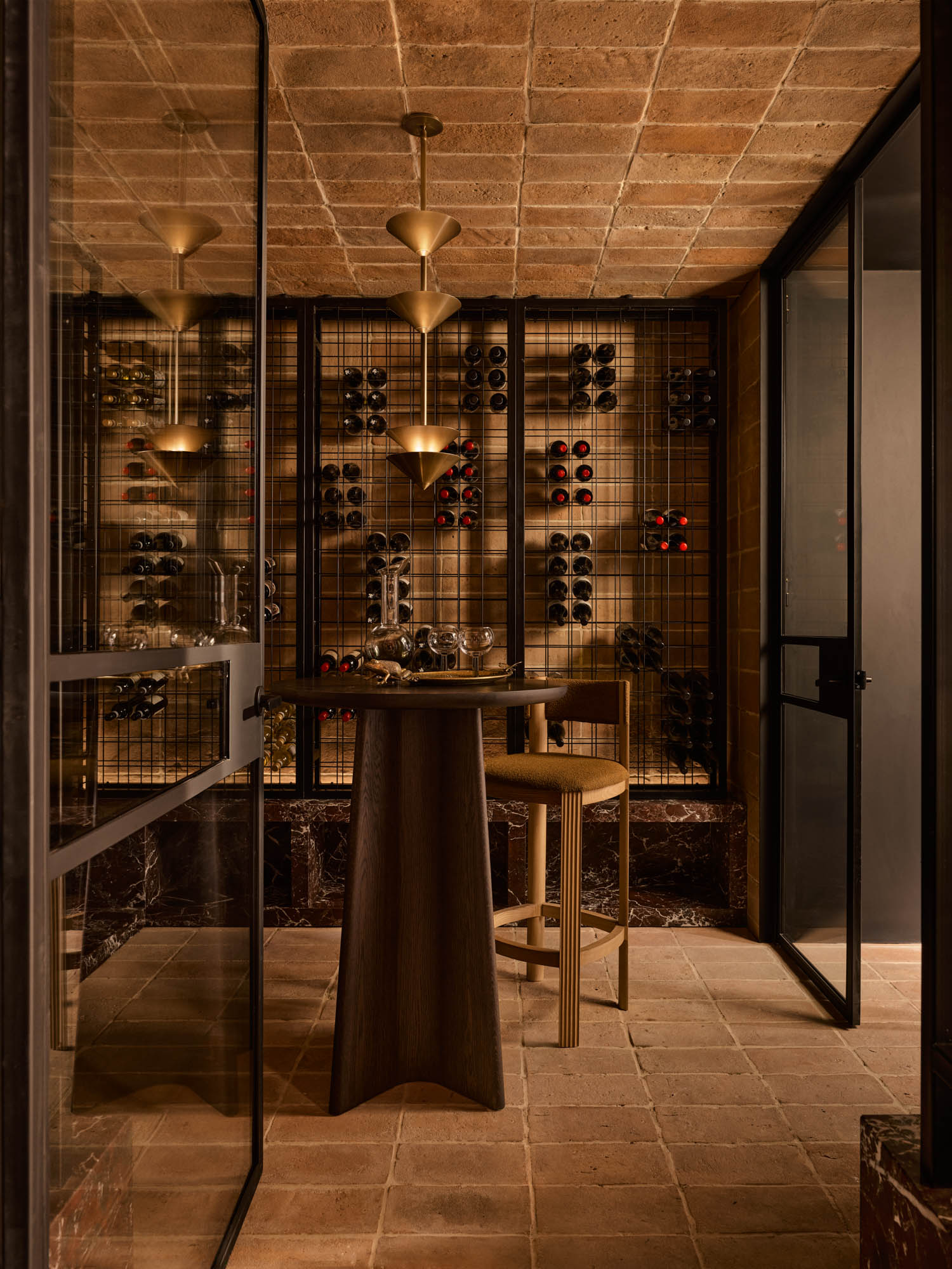 A modern wine cellar with glass doors, a wooden table and stool, grid shelves holding wine bottles, and warm pendant lighting.
