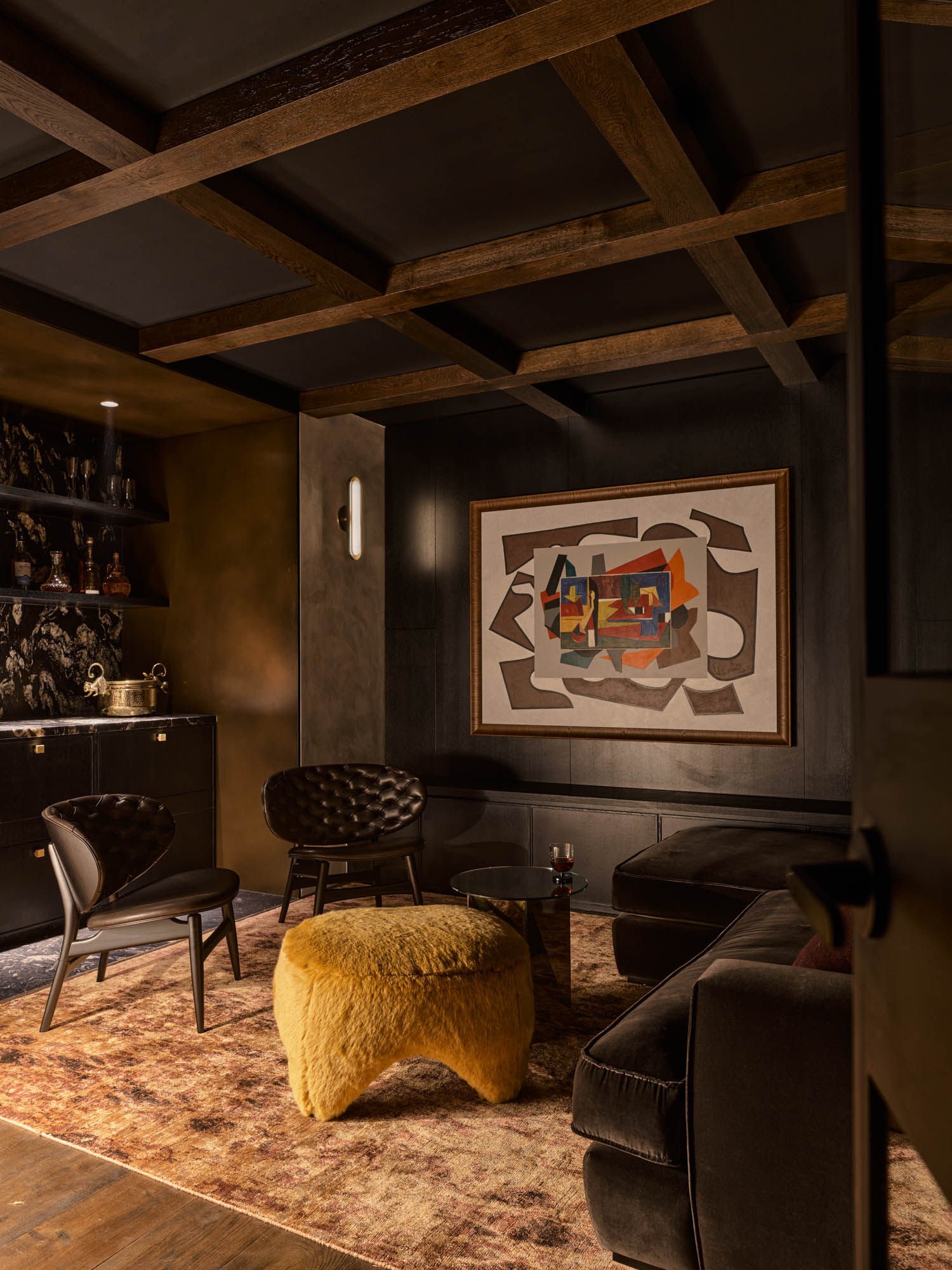 A dimly lit lounge features dark walls, modern chairs, a yellow fuzzy ottoman, abstract wall art, and shelves with decor.