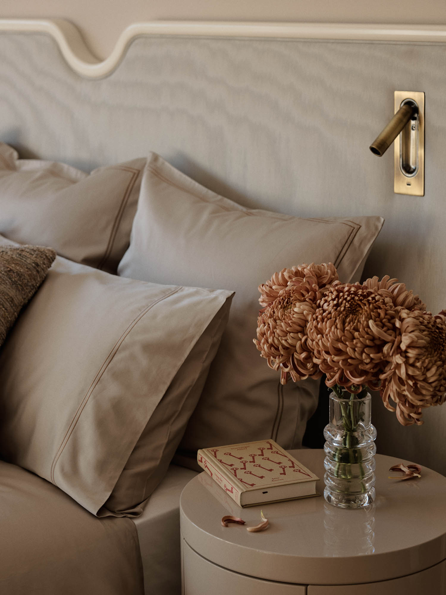A bedside table with a glass vase of dried flowers, a closed book, and a bed with beige bedding and pillows against a light wood headboard.