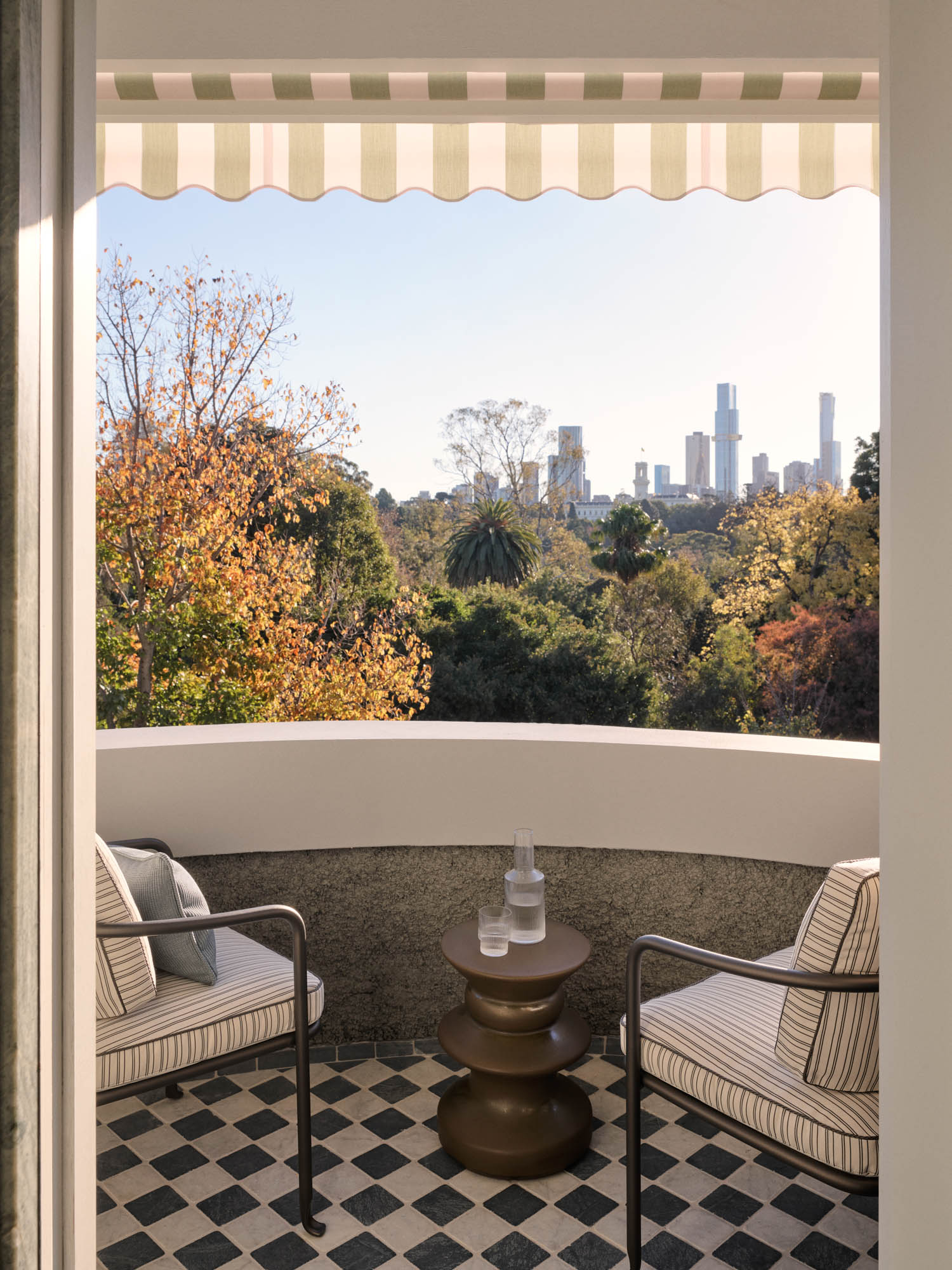 Two striped patio chairs and a small table with a glass and water carafe sit on a checkered balcony, overlooking trees and a distant city skyline under a striped canopy.