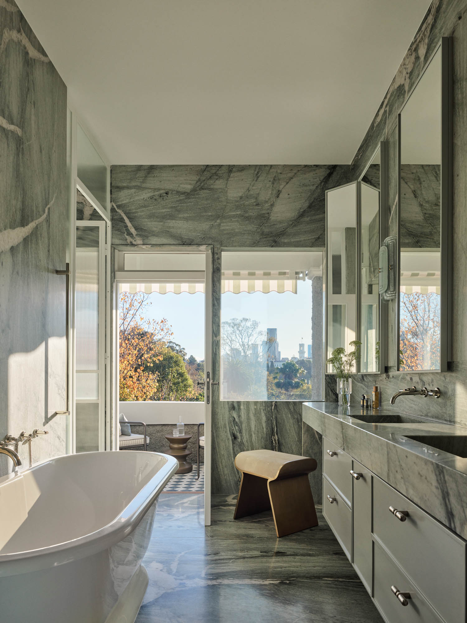 Bathroom with marble walls and floor, freestanding bathtub, double vanity, and large windows letting in natural light, with a view of trees and buildings outside.