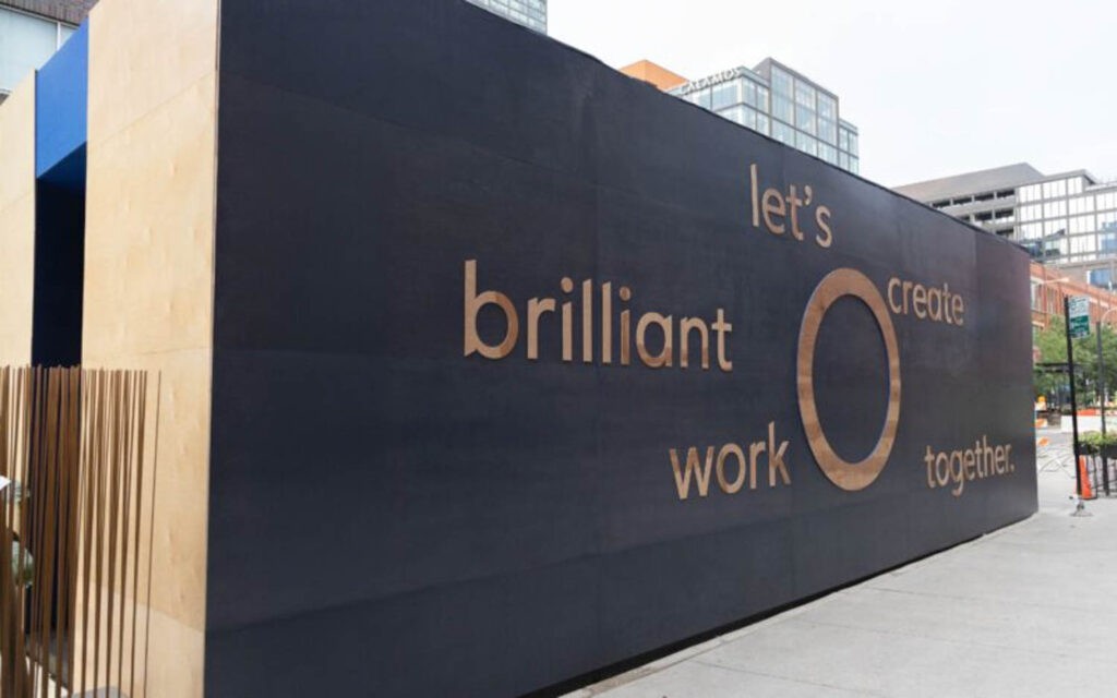A black wall with wooden letters reads, "let’s create brilliant work together," on a city sidewalk with buildings in the background.
