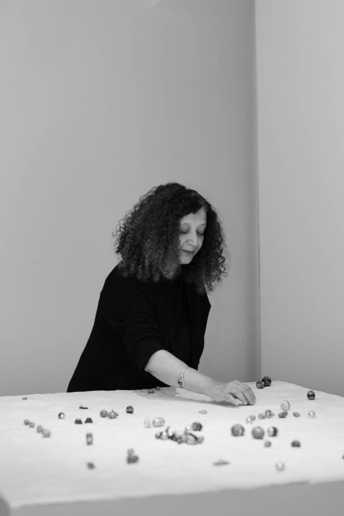 A woman with curly hair arranges small objects on a white table in a minimalist room. The image is in black and white.