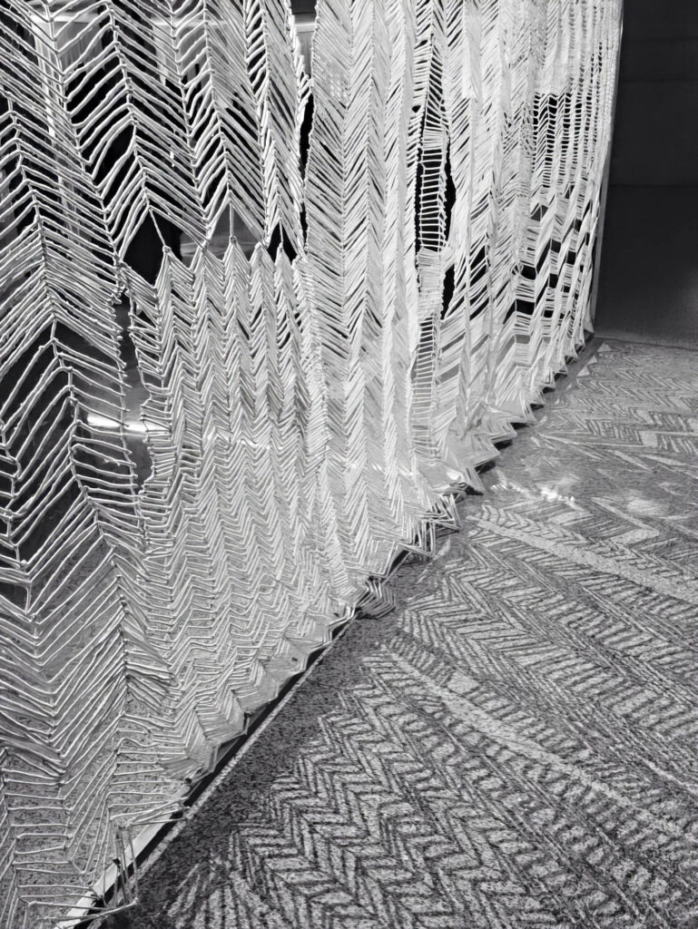 A geometric metal sculpture with repeating chevron patterns stands on a textured floor with similar herringbone designs, both in black and white tones.