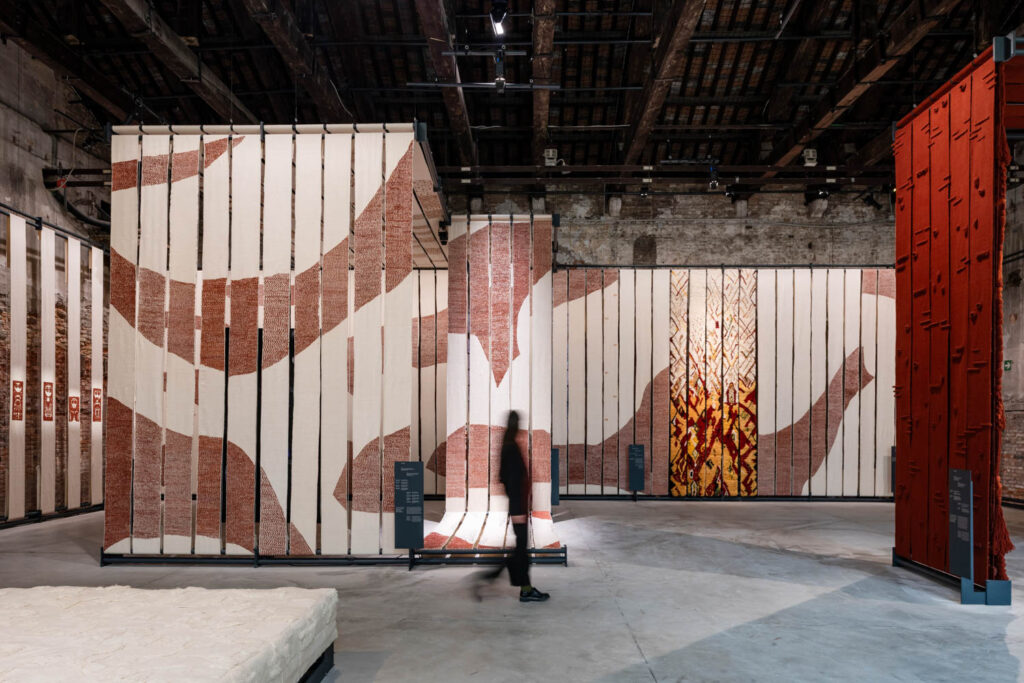A person walks through an art exhibit featuring large textile panels with abstract patterns in a spacious, industrial-style gallery.