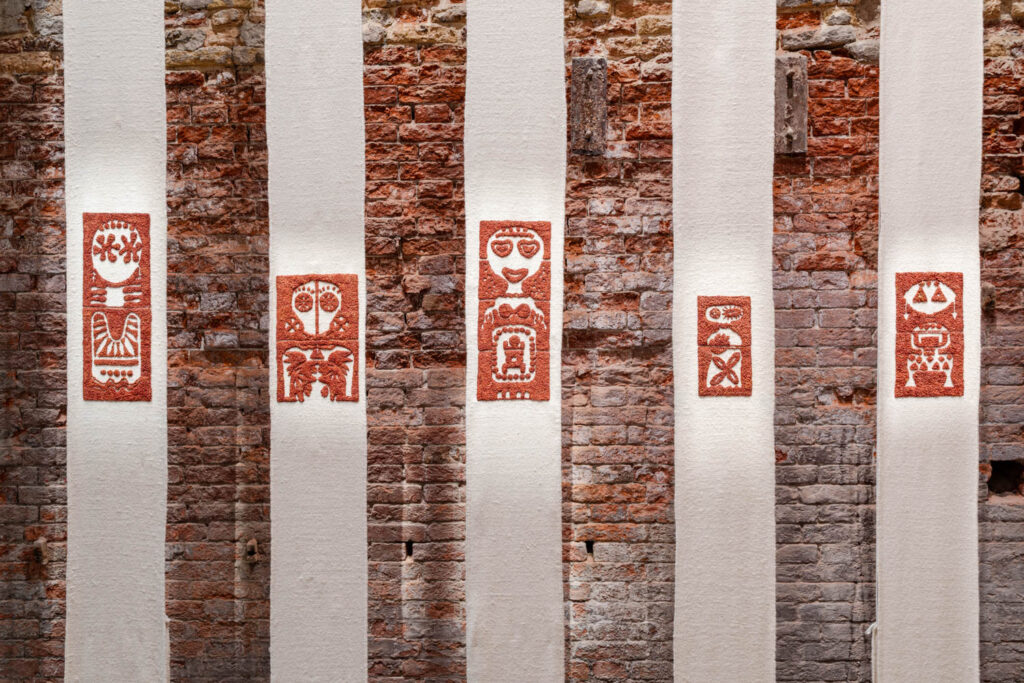 Rectangular textile artworks with red symbols are displayed vertically on white fabric panels against a rustic brick wall.