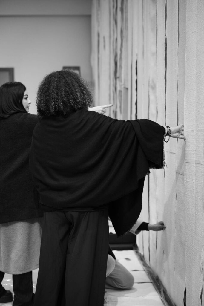Two people stand side by side indoors, one with an arm outstretched toward a wall covered with vertical fabric strips. The scene is captured in black and white.
