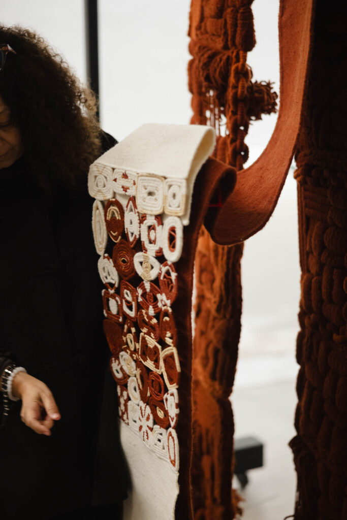 A person holds a textile with intricate red and white patterns next to a larger, textured reddish-brown fabric.
