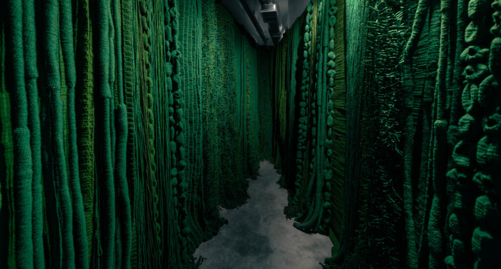 A narrow hallway with walls covered in thick, textured green fabric resembling dense foliage, and a concrete floor.