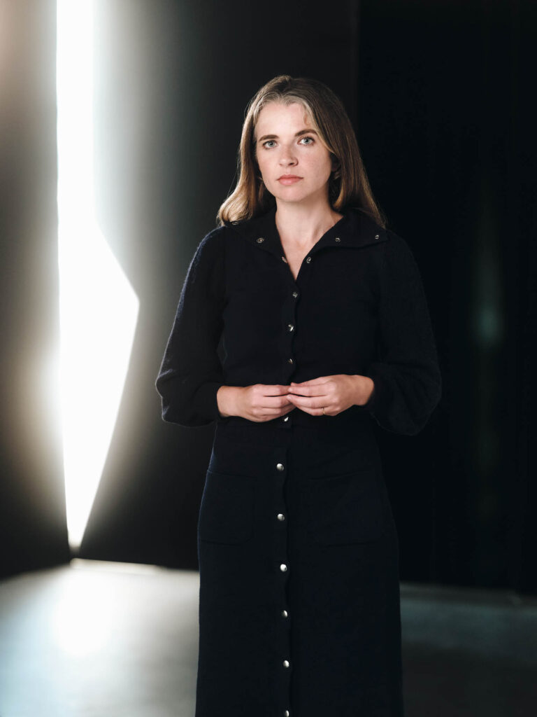 Woman in a dark buttoned dress stands indoors with hands clasped, facing the camera, with a bright vertical light in the background.