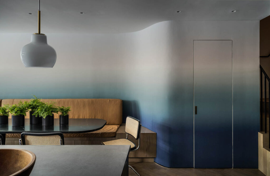 Modern dining area with a curved wall featuring a blue gradient, a tan bench, black table, potted plants, woven chairs, and a white pendant light.