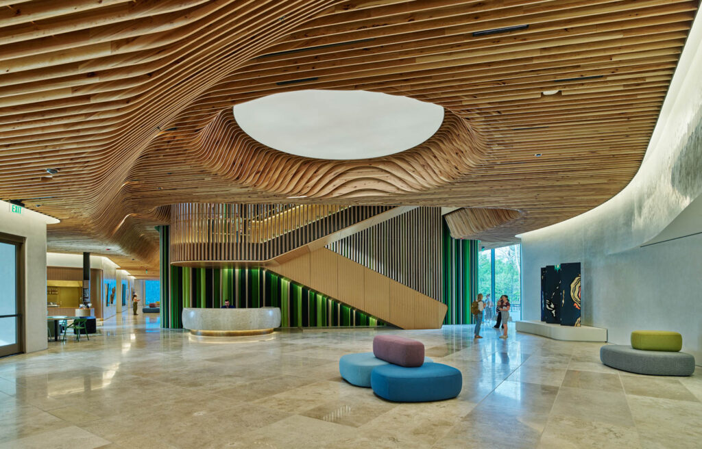 Modern lobby with curved wooden ceiling, central circular skylight, marble floors, colorful seating, reception desk, and a staircase. Three people stand near the staircase.