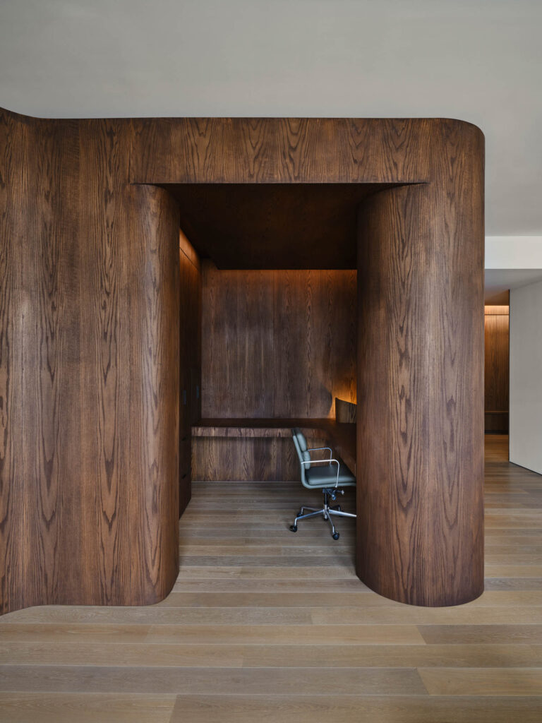 Curved dark wood alcove with a built-in desk and a gray office chair on light wood flooring in a modern interior space.