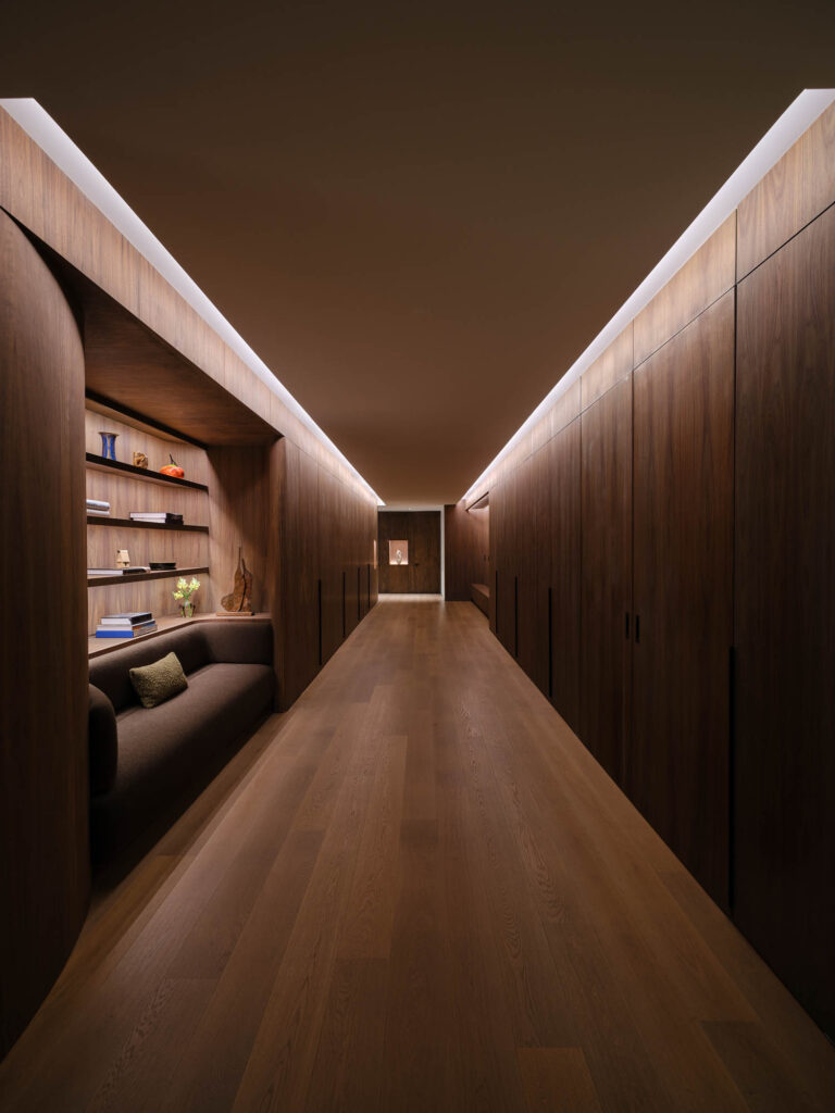 A modern hallway with wood paneling, built-in shelves, a small sofa, minimal decor, and recessed ceiling lighting.