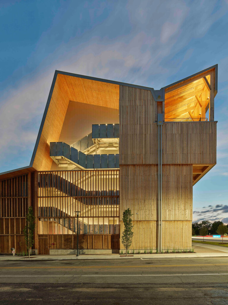 Modern building with wooden exterior, open-air staircases, and angular roof, photographed at dusk with soft lighting and a partly cloudy sky in the background.