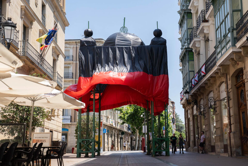 A large black and red fabric structure covers a street installation in a European city, surrounded by outdoor café seating and historic buildings.