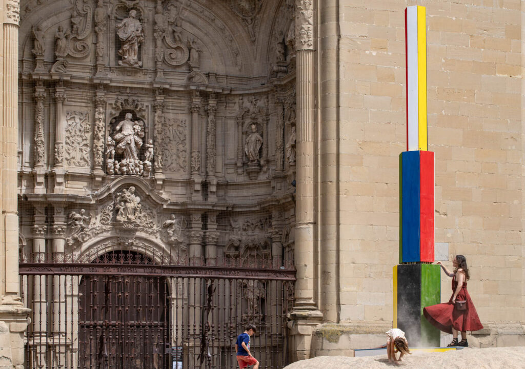 A woman stands beside a tall, colorful rectangular sculpture in front of an ornate stone building, while two children play nearby.