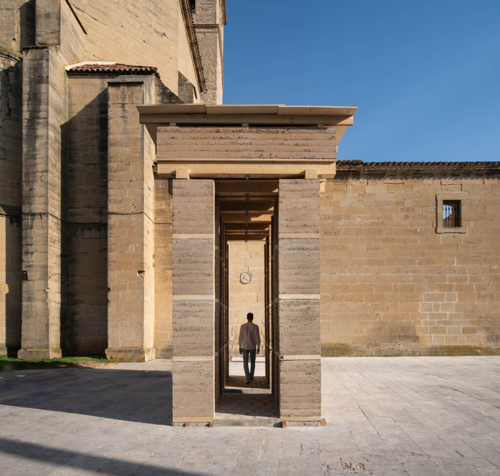A person stands inside a rectangular stone structure with columns, positioned in front of a large beige stone building under a clear blue sky.