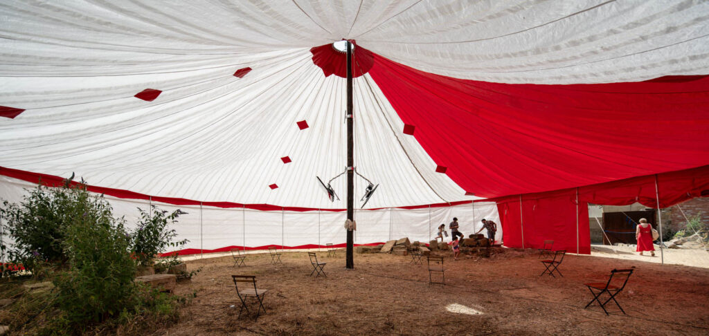 Interior of a red and white circus tent with a dirt floor, scattered empty chairs, and several people standing near the open entrance.
