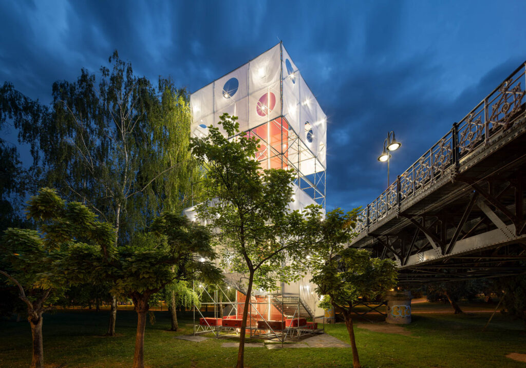 A tall, illuminated open-frame tower with geometric banners stands near trees and a metal bridge at dusk.