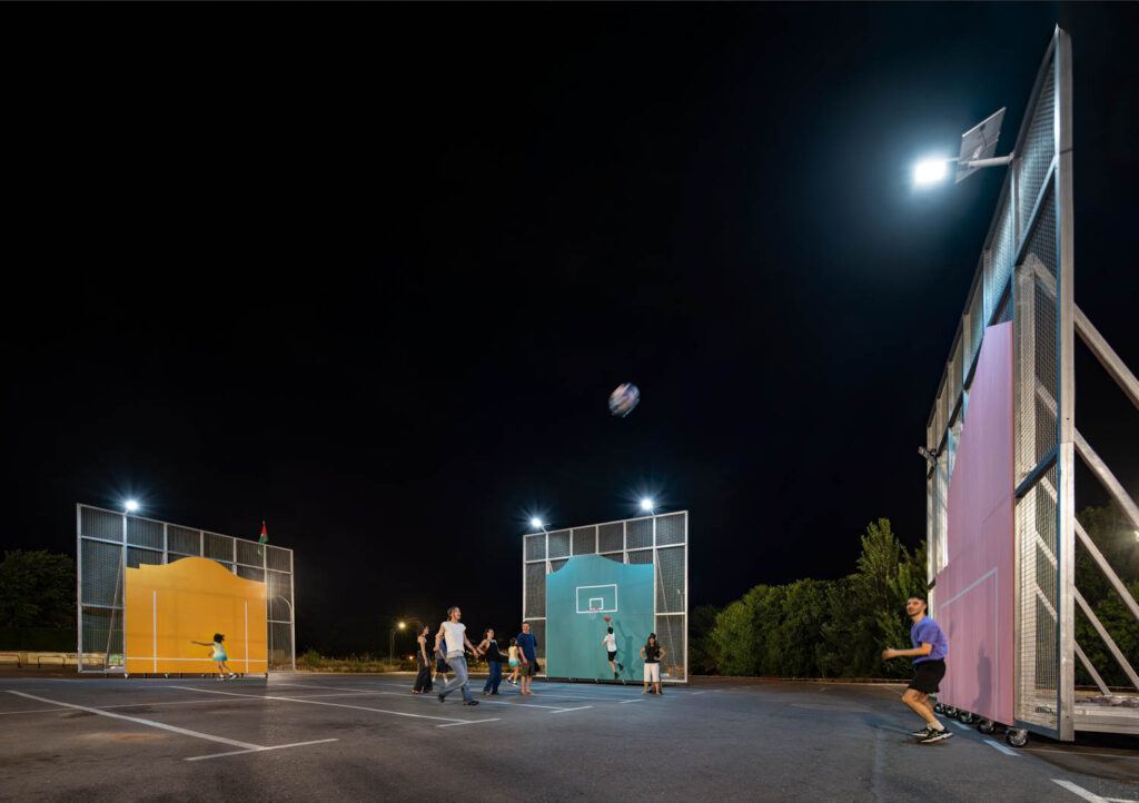 A group of people play basketball at night on an outdoor court with colorful backboards and overhead lights.