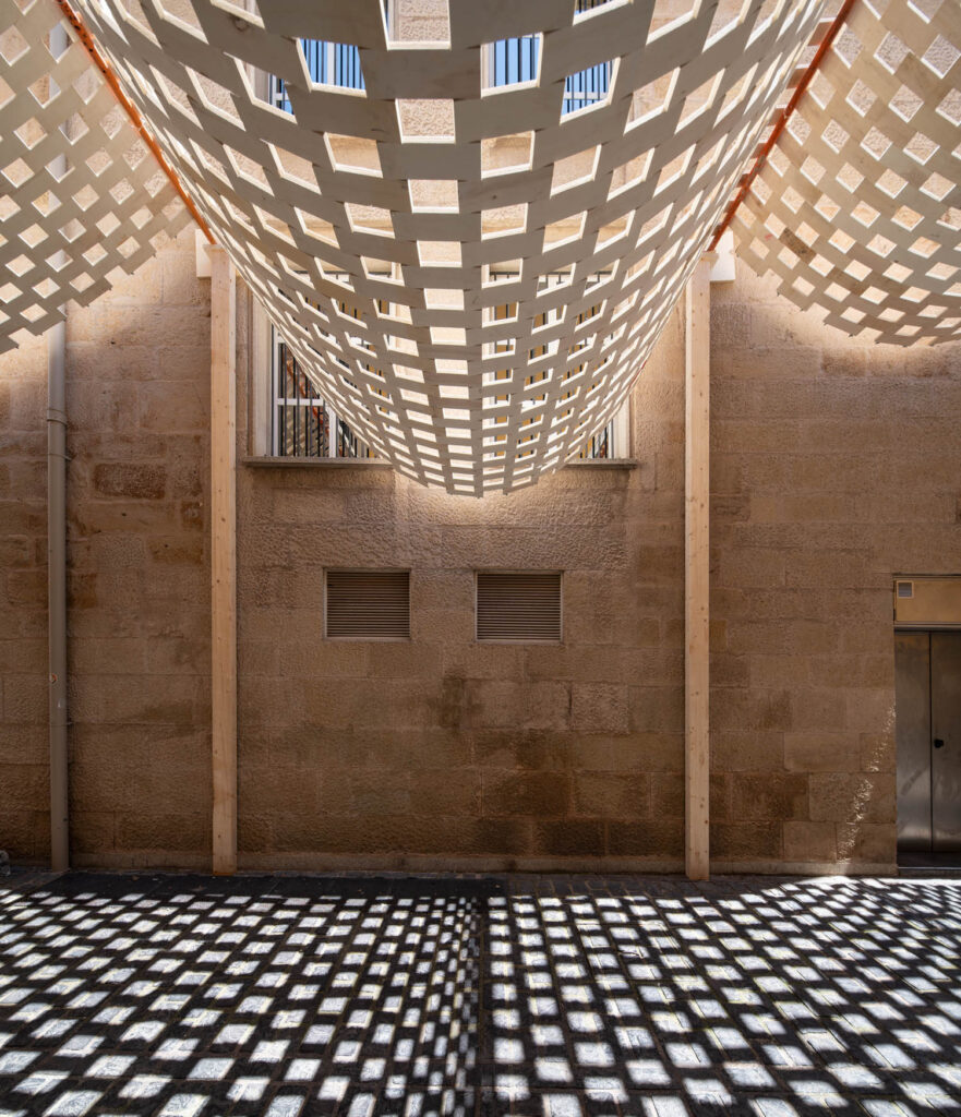 A lattice canopy suspended between stone walls casts a geometric shadow pattern on the ground below.