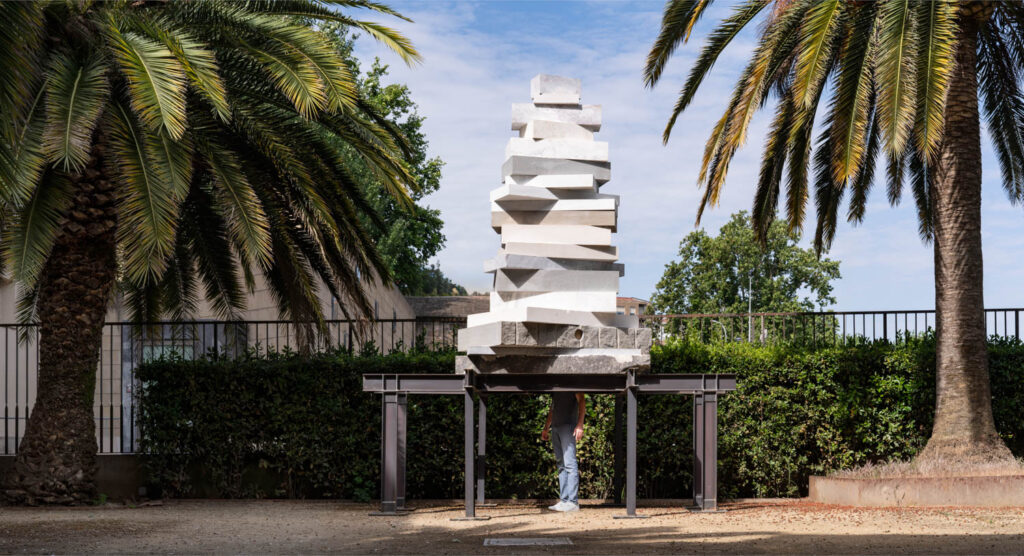 A person stands under a large outdoor sculpture made of stacked, irregular white stone slabs supported by a metal frame, with palm trees and bushes in the background.