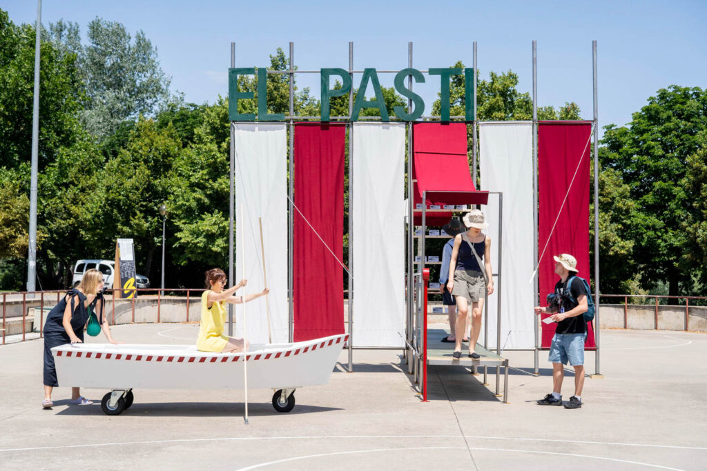 Four people interact with a theatrical outdoor setup featuring a small white boat and a structure with red and white panels labeled "EL PASTI." Trees and blue sky are in the background.