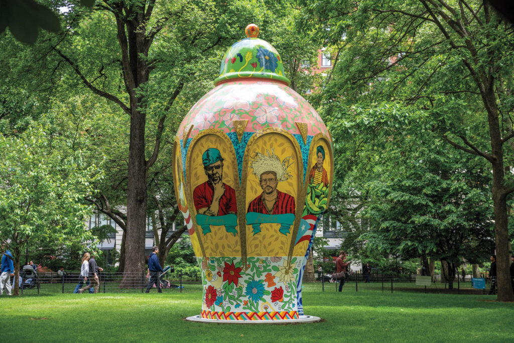Large, colorful urn-shaped sculpture with painted portraits stands in a green park surrounded by trees, with people walking nearby.