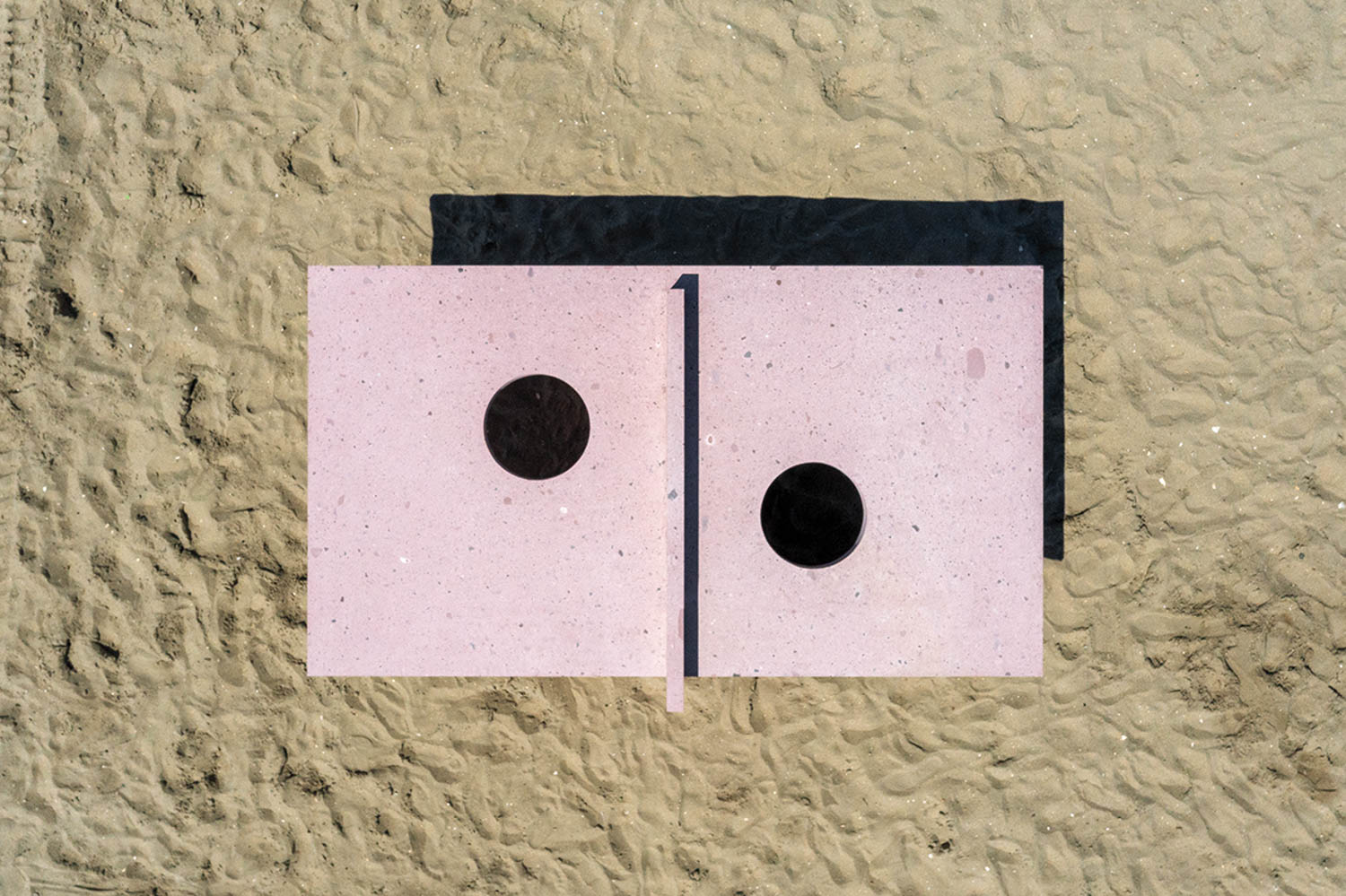 Aerial view of two pink rectangular slabs with round holes, placed side by side on sand, casting a shadow behind them.