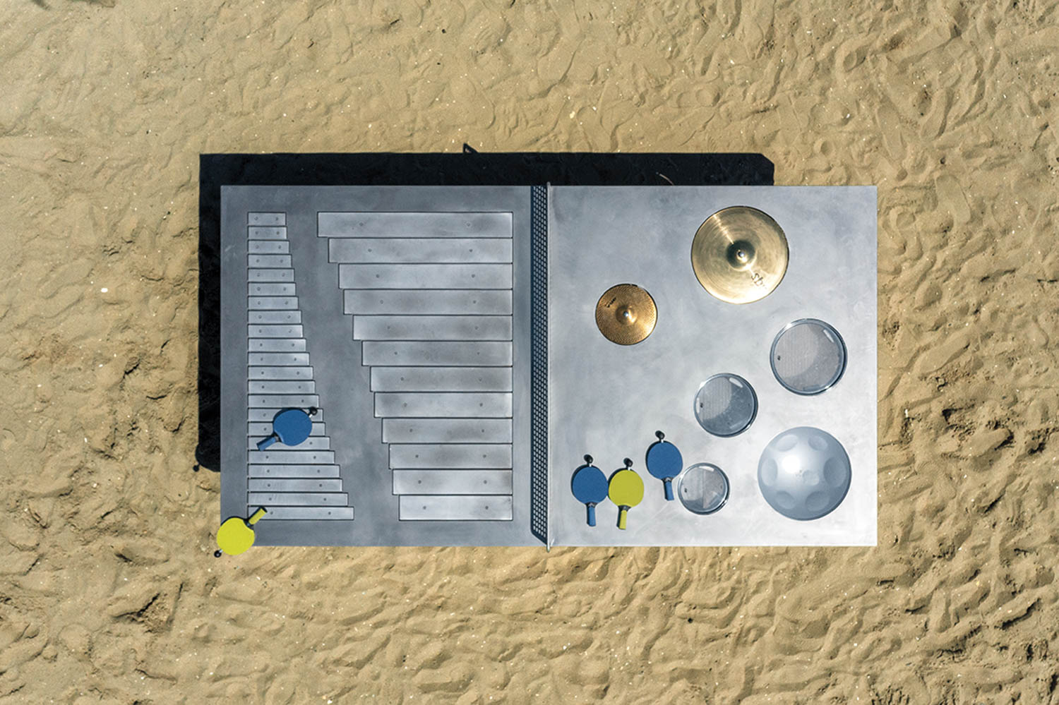 Overhead view of a metallic outdoor musical instrument panel with percussion disks, cymbals, and mallets, installed on sandy ground.