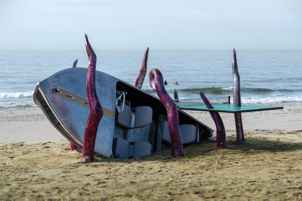 An overturned rowboat on the beach is surrounded by large, sculpted purple tentacles appearing to rise from the sand, with the ocean in the background.
