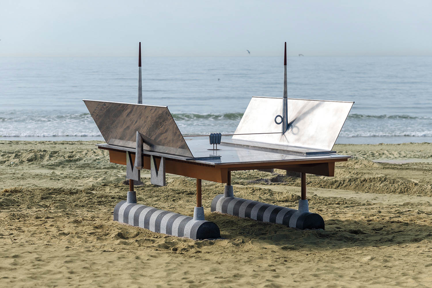 A table tennis table with paddles and net is placed on a sandy beach near the ocean, featuring unusual sculptural elements on its legs and sides.