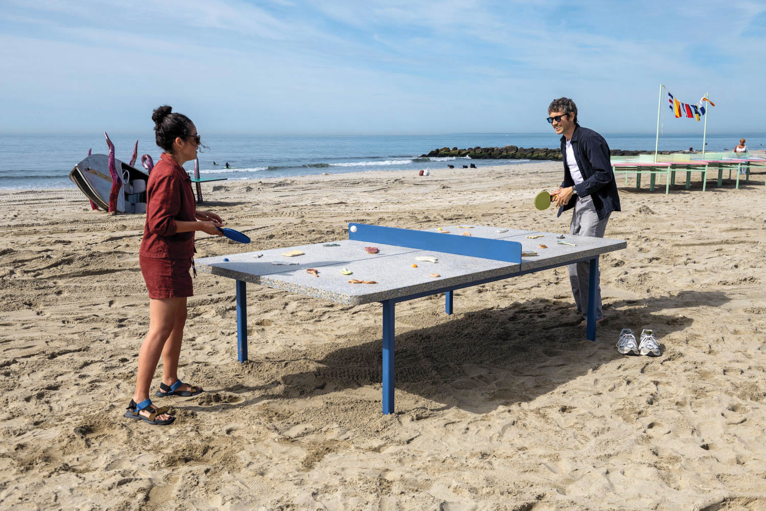 Two people play table tennis on a sandy beach with food scattered on the table, ocean and boats in the background, and flags displayed on poles nearby.