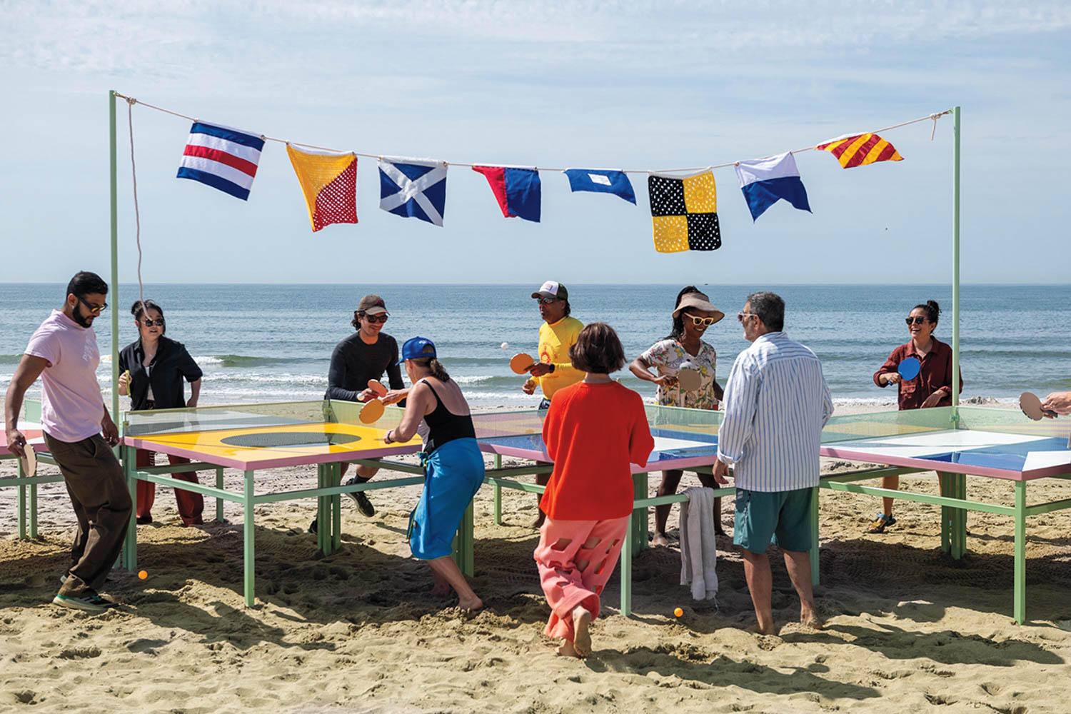 A group of people play table tennis on a beach around a large multi-sided table, with colorful flags strung above and the ocean in the background.