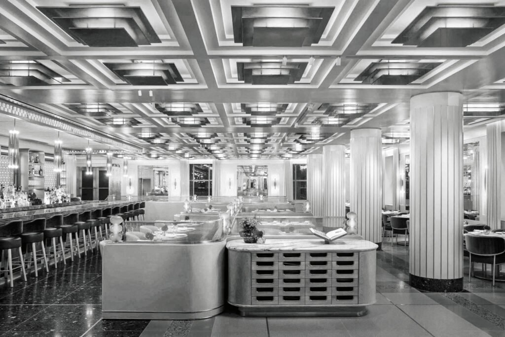 Black and white photo of a spacious, art deco style restaurant with a geometric ceiling, columns, long bar, and booth seating. Tables are set and lighting fixtures line the ceiling and walls.