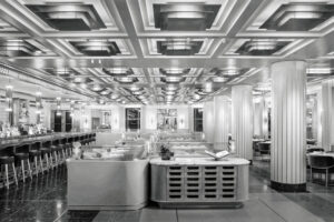 Black and white photo of a spacious, art deco style restaurant with a geometric ceiling, columns, long bar, and booth seating. Tables are set and lighting fixtures line the ceiling and walls.