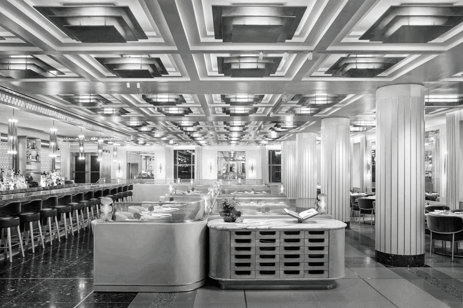 Black and white photo of a spacious, art deco style restaurant with a geometric ceiling, columns, long bar, and booth seating. Tables are set and lighting fixtures line the ceiling and walls.