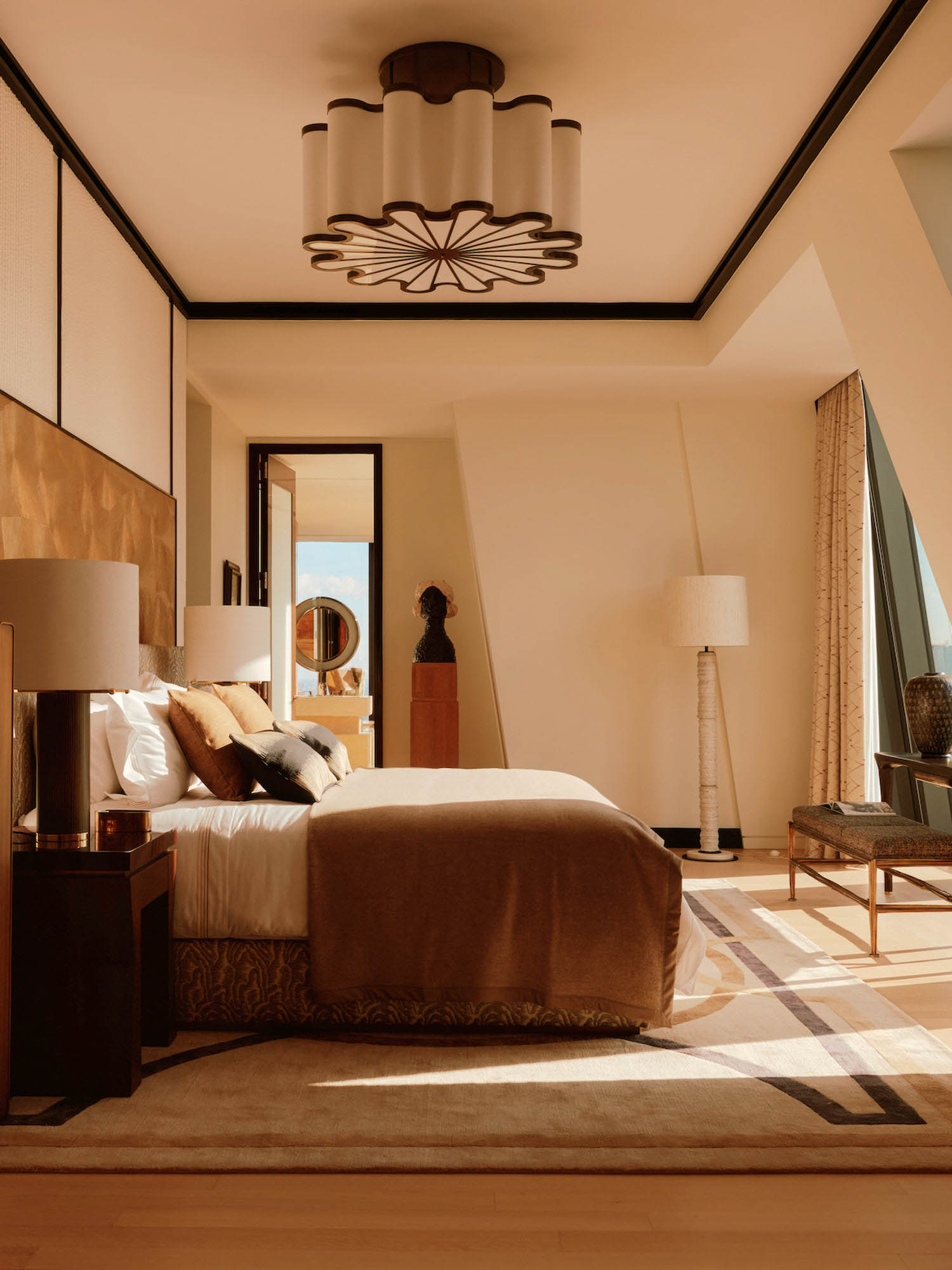 Modern bedroom with a large bed, neutral bedding, geometric rug, tall lamp, accent chair, and a sculptural ceiling light, all in warm tones with natural light from a window.