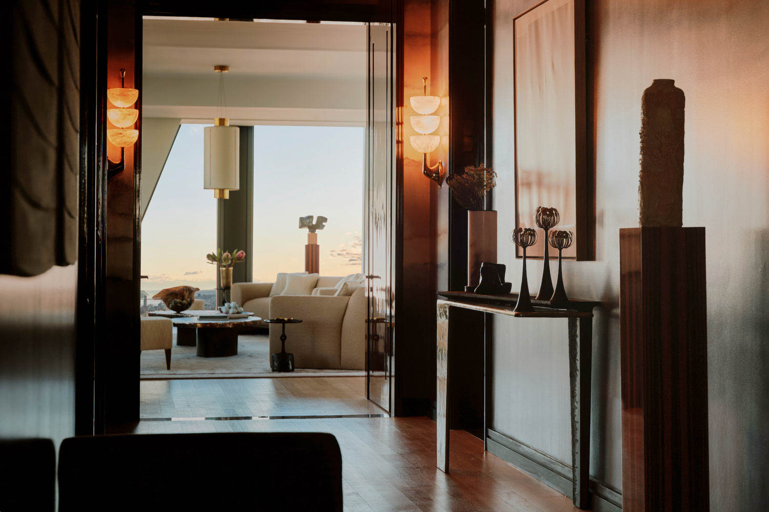 A hallway with wall sconces leads to a living room with beige sofas, large windows, and a view of the sky outside. Modern decor and ambient lighting create a warm atmosphere.