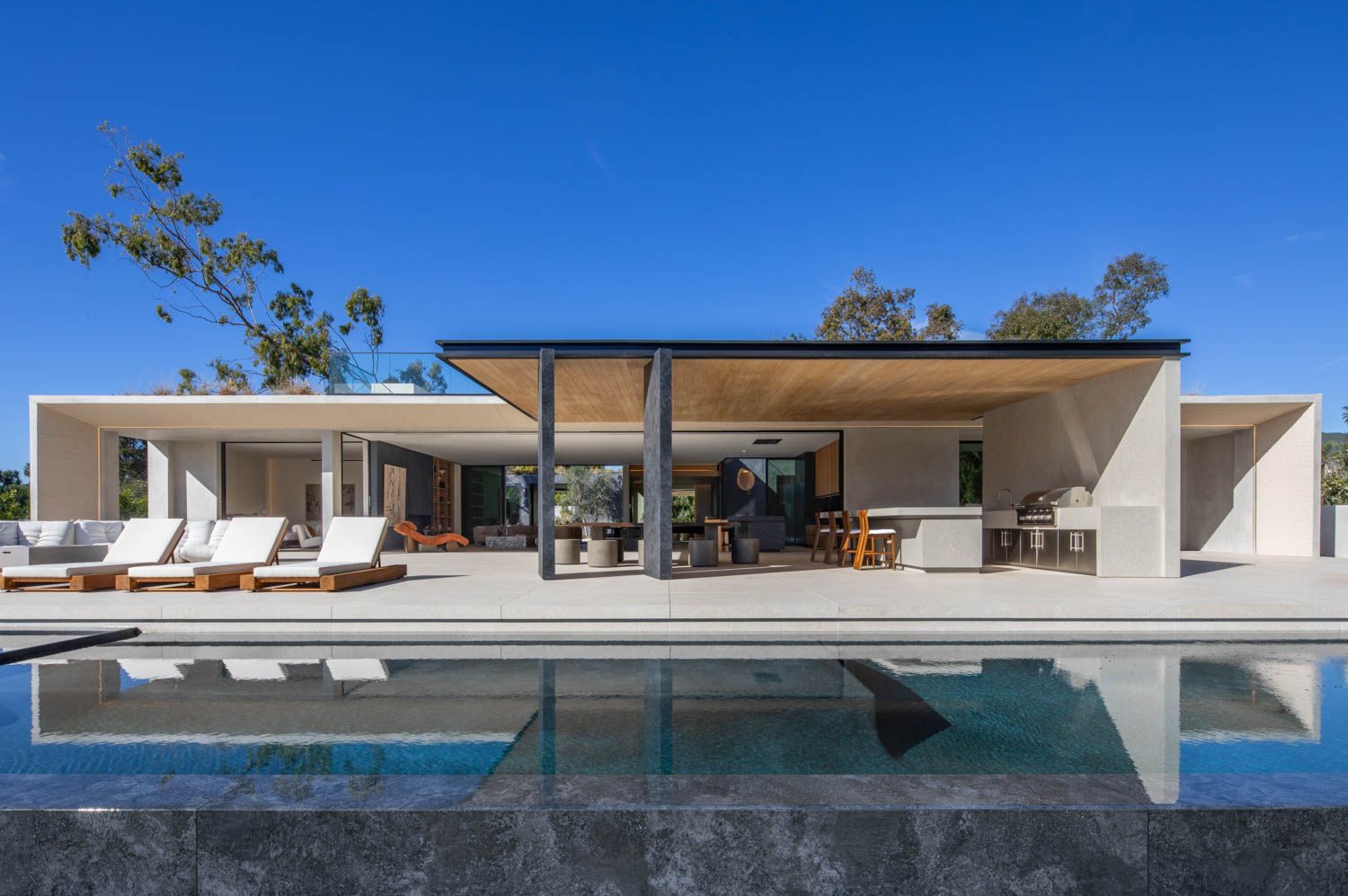Modern minimalist house with open patio, outdoor seating, barbecue area, and a swimming pool under clear blue sky.
